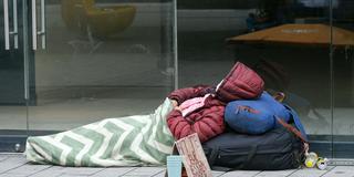 Eine obdachlose Person liegt mit Jacke und Decke vor einem leerstehenden Ladenlokal auf dem Gehweg (Foto: IMAGO / Michael Gstettenbauer)