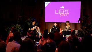 Blick vom Publikum auf die Bühne, dort sitzen drei Personen. Im Hintergrund steht auf einer lila Wand "Liebt Euch" geschrieben. (Foto: Liebt Euch Podcast)