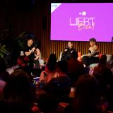 Blick vom Publikum auf die Bühne, dort sitzen drei Personen. Im Hintergrund steht auf einer lila Wand "Liebt Euch" geschrieben. (Foto: Liebt Euch Podcast)