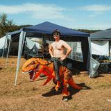 Ein Festivalbesucher steht oberkörperfrei auf dem Campingplatz, er trägt ein Kostüm, das aussieht, als würde er auf einem Dino reiten. (Foto: MxM Photo)