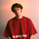 Ein junger Mann mit mittellangen braunen Haaren steht vor einem hellen Hintergrund. Er trägt ein rotes T-Shirt und guckt mit neutralem Gesichtsausdruck in die Kamera. (Foto: Pressefoto/VIOSIONV)